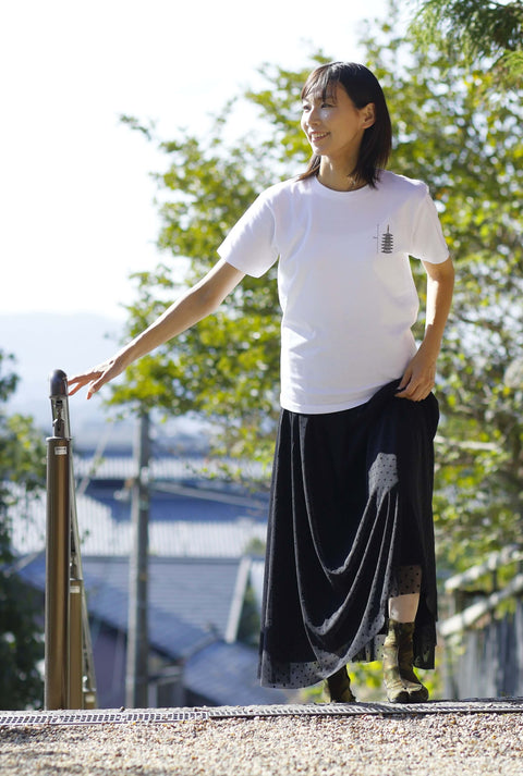 Woman wearing a white T-shirt named “Five-story pagoda & Kaiju Monster”, featuring a printed design of a traditional Japanese pagoda with a small kaiju monster beside it. She is outdoors, holding a handrail, and wearing a long black skirt with a dotted pattern.