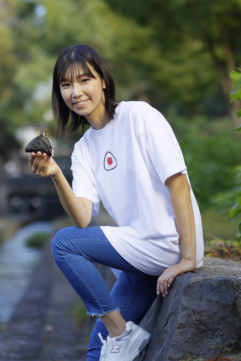 Woman wearing a white T-shirt named “Onigiri”, featuring a printed design of a rice ball with a red umeboshi center on the chest. She is outdoors, smiling, and holding a real onigiri while sitting on a stone ledge near a stream.