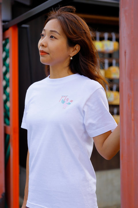 Woman wearing a white T-shirt named “Dove”, featuring a soft-colored printed design of a torii gate and a dove on the chest. She is standing in front of a traditional Japanese setting, gently touching a red pillar.