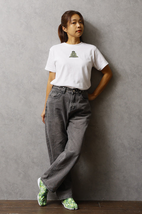 Woman wearing a white T-shirt named “Kamakura Daibutsu”, featuring a printed design of the Great Buddha of Kamakura on the chest. She is standing against a gray wall, with one leg crossed and hands relaxed.