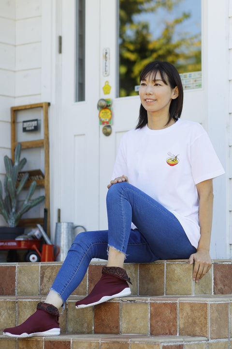 Woman wearing a white T-shirt named “Ramen”, featuring a colorful printed design of a bowl of ramen with chopsticks on the chest. She is sitting on tiled steps in front of a white door, wearing burgundy slip-on shoes and blue jeans.