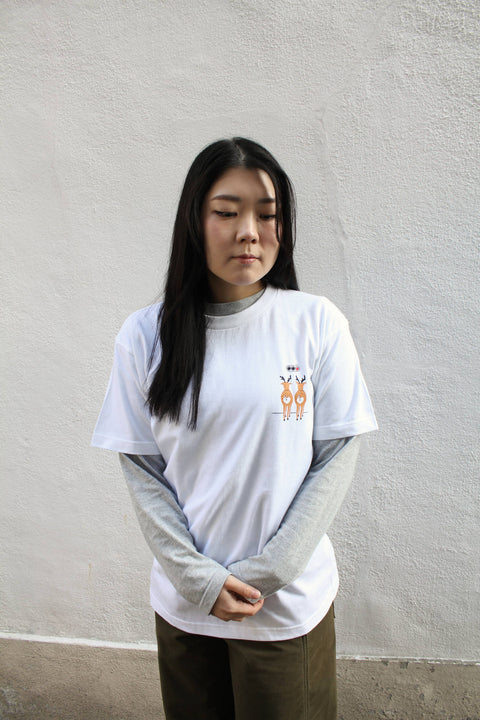 Layered outfit featuring a gray long-sleeved T-shirt worn under a white T-shirt with the “Deer Walk” design, standing against a white wall